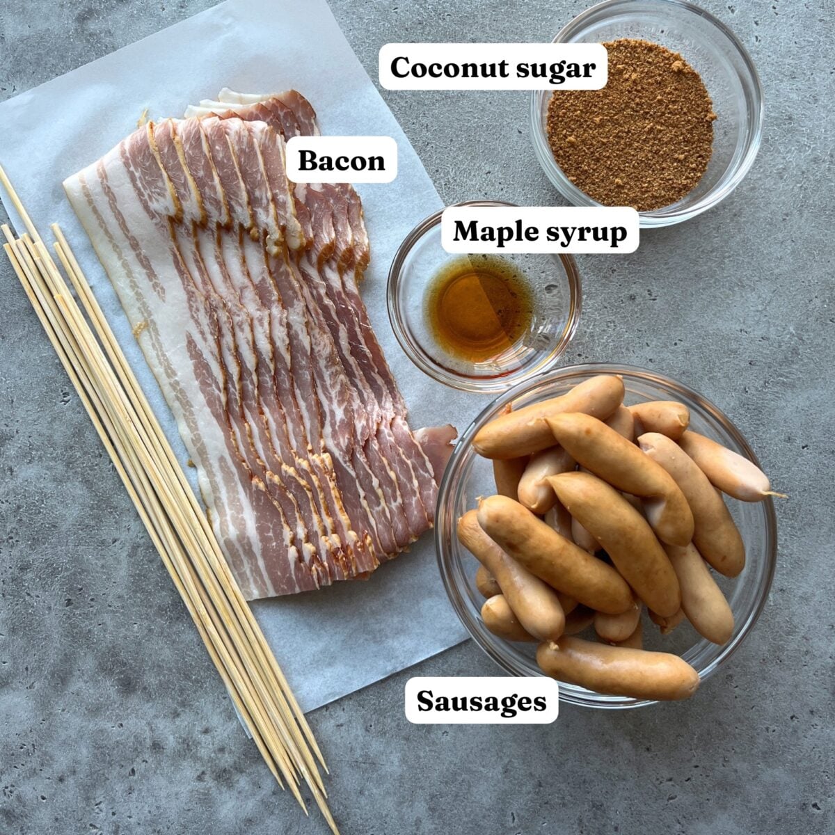 Bacon slices, mini sausages, wooden skewers, coconut sugar, and a small bowl of maple syrup arranged on a gray surface.