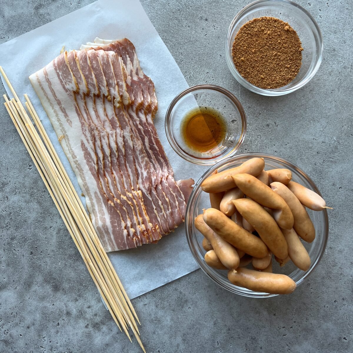 Sliced bacon, small sausages, wooden skewers, coconut sugar, and a small bowl of maple syrup arranged on a gray surface.
