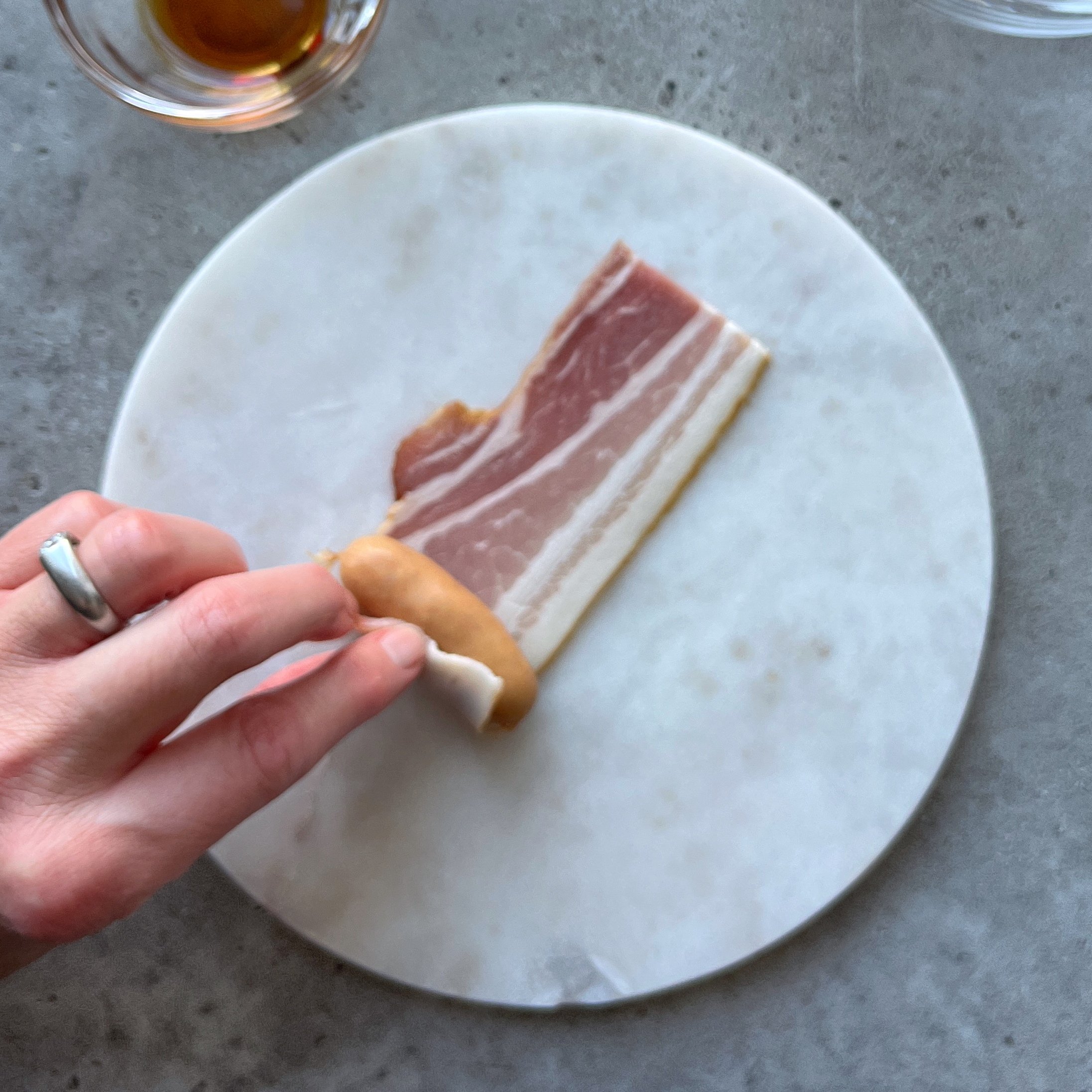 A hand rolls a sausage in a slice of raw bacon on a round white cutting board, with a small bowl of liquid nearby.