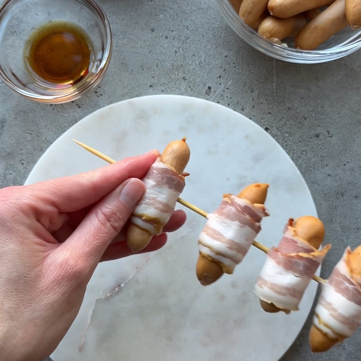 A hand holds a skewer with small sausages wrapped in bacon, positioned over a round white plate on a gray surface. A bowl of maple syrup and a bowl of sausages are nearby.