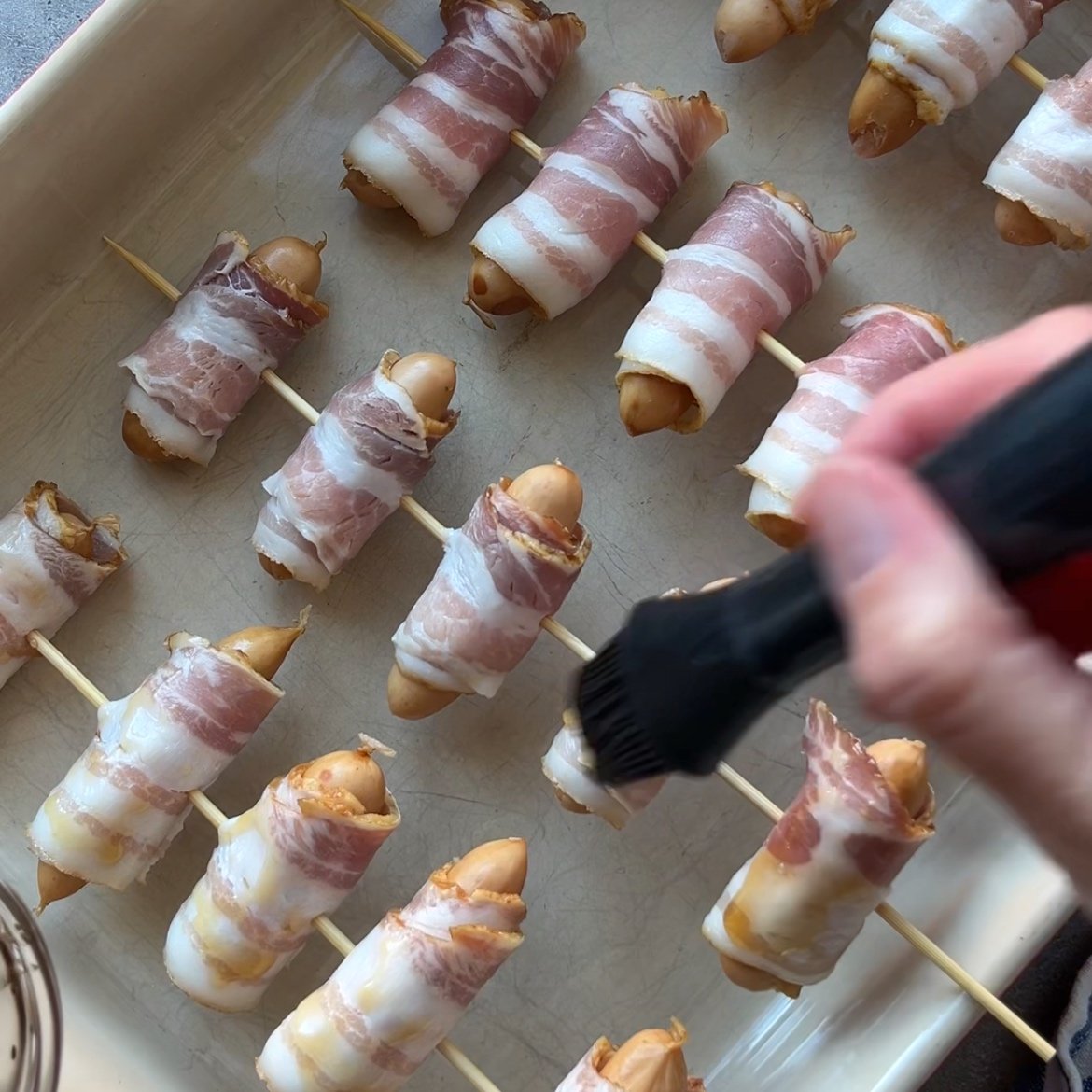 A hand brushes a maple glaze onto bacon-wrapped sausages arranged on skewers in a baking dish.