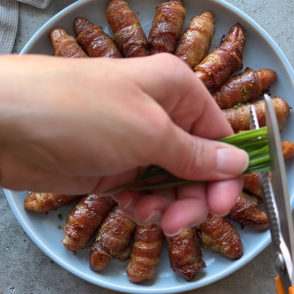 A hand uses scissors to cut chives over a plate of bacon-wrapped snacks arranged in a circular pattern.