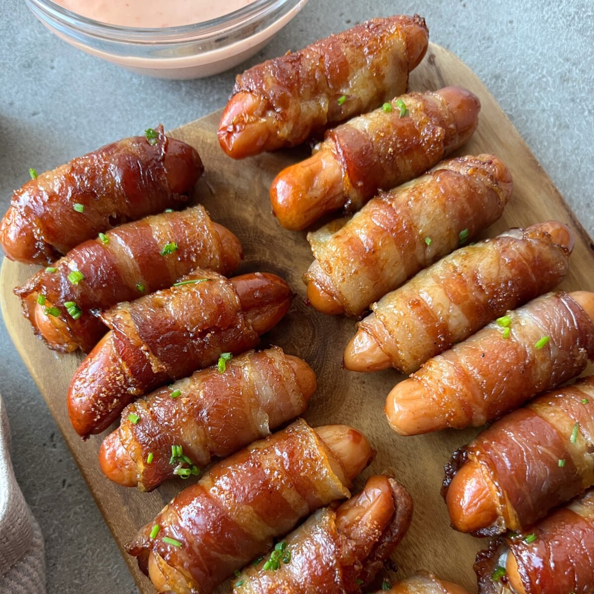 A wooden board with several bacon-wrapped sausages, garnished with chopped herbs, next to a bowl of dipping sauce.