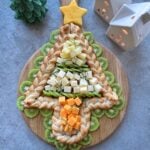 A Christmas tree-shaped pastry braid topped with cheese cubes and surrounded by kiwi slices on a wooden board, with decorative ceramic houses in the background.