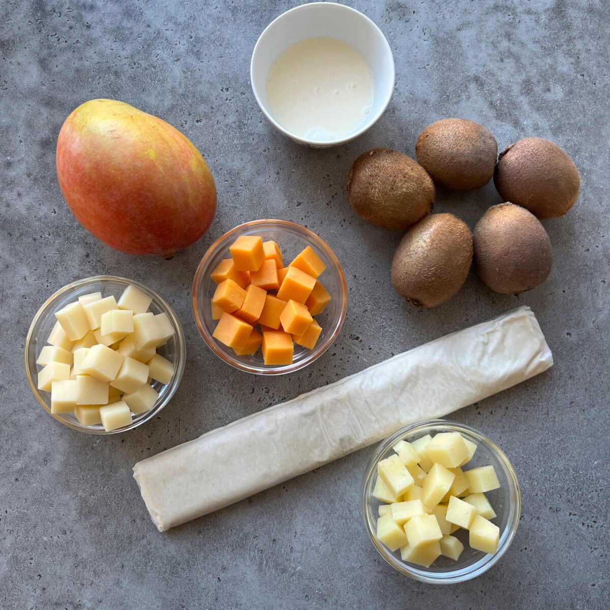 A mango, four kiwis, a bowl of cream, three bowls of cubed cheese, and a sheet of pastry are arranged beautifully on a gray surface.