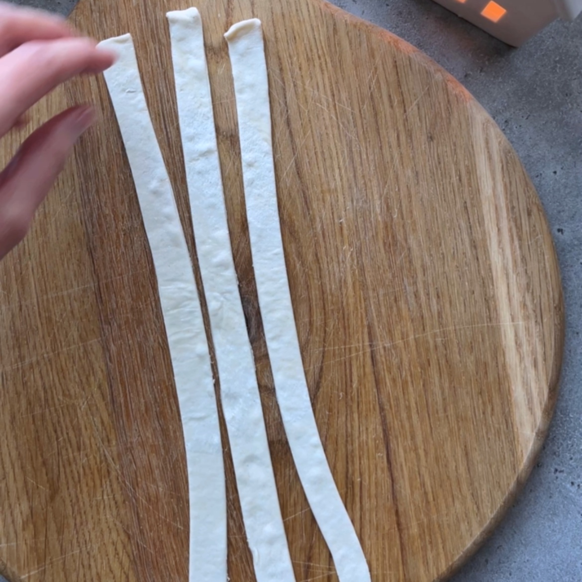Three uncooked dough strips are laid out parallel on a round wooden cheese board, with a hand reaching toward them from the left side of the image.