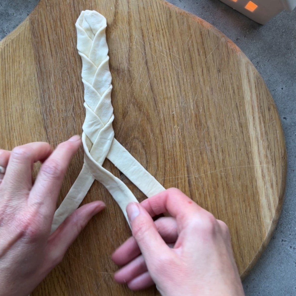 Two hands braiding three strips of dough on a round wooden board.