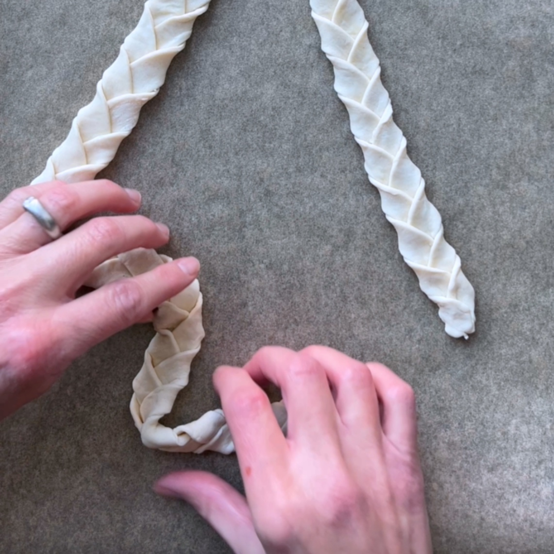Two braided strips of dough are on parchment paper; a person’s hands are shaping one of the strips into a tree.