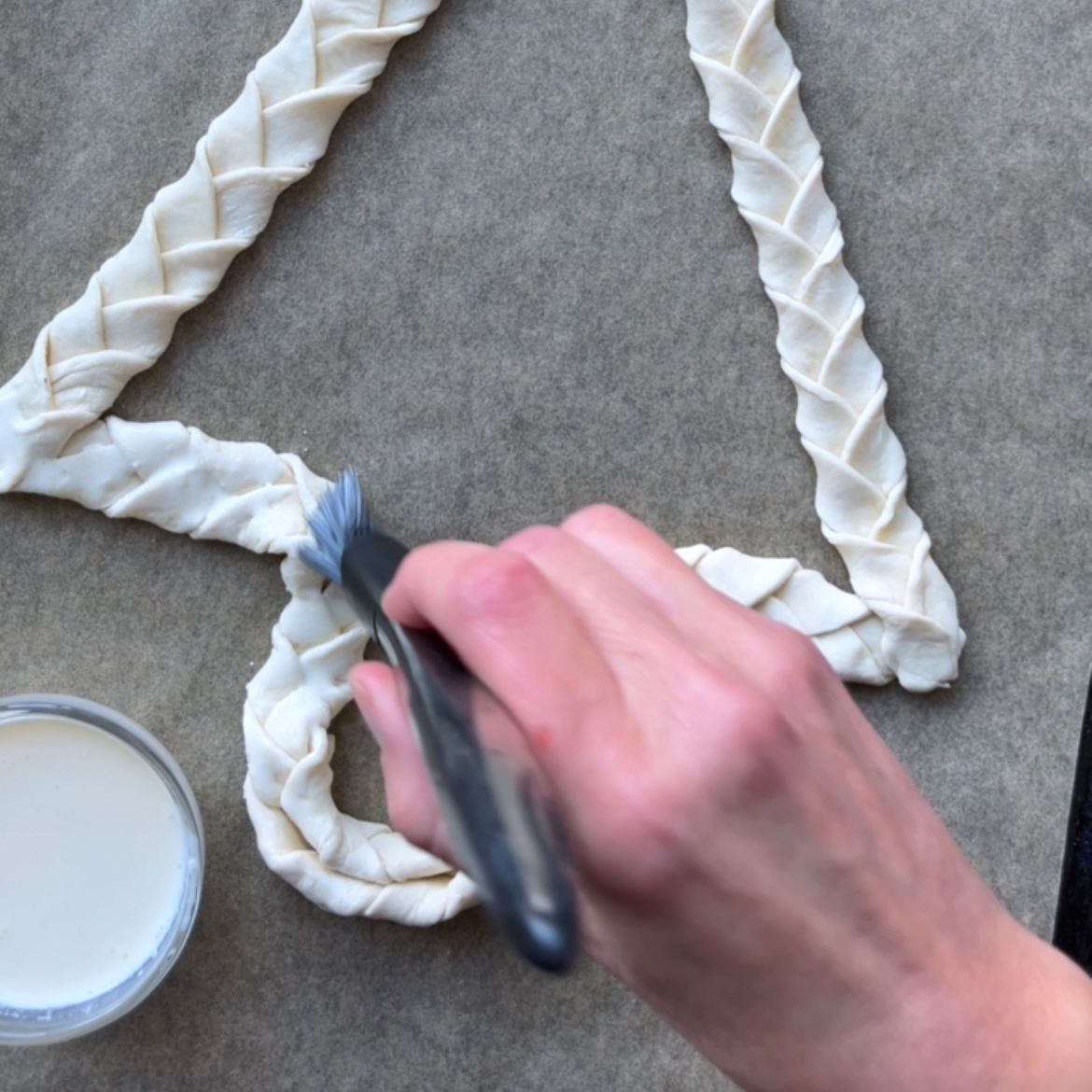 A hand uses a pastry brush to apply heavy cream to braided dough shaped like a Cheese Board Christmas Tree on parchment paper.