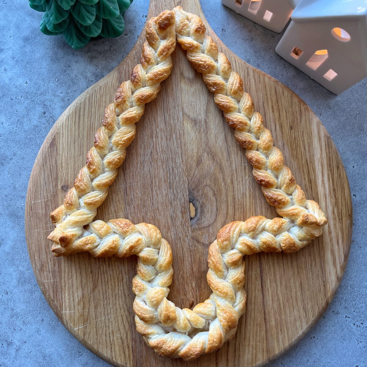 A braided pastry shaped like a Christmas tree sits on a round wooden cheese board; ceramic houses and a green tree decoration are nearby.