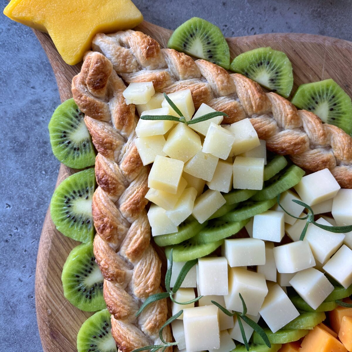 A cheese board is arranged decoratively with sliced kiwi, cubed white cheese, braided puff pastry, and a star-shaped cheese piece on top.