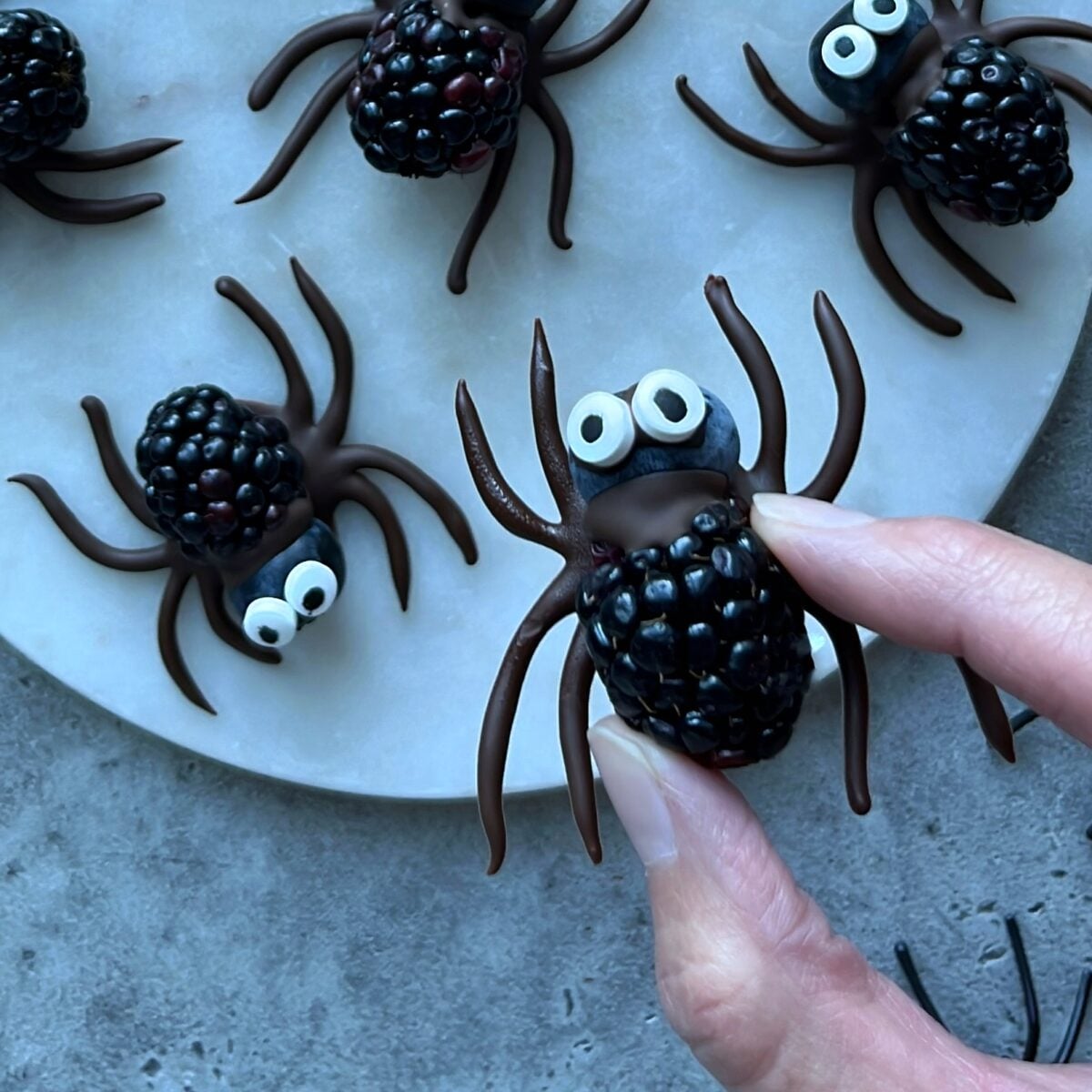 Hand holding a blackberry decorated as a spider with a blueberry head, chocolate legs and candy eyes, with similar blackberry spiders arranged on a white plate.