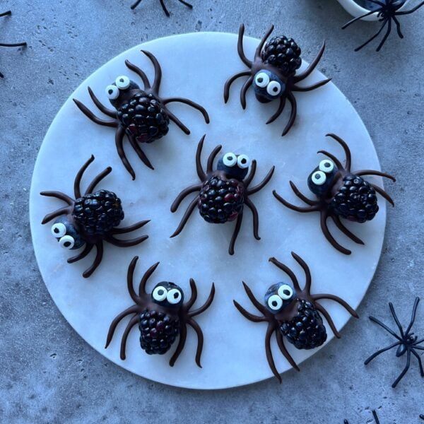 Seven spider-shaped treats made from blackberries, blueberries, chocolate, and candy eyes are arranged on a round white plate.