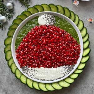A plate arranged with pomegranate seeds, pesto, and whipped feta dip in the shape of a Santa hat, surrounded by sliced cucumbers.