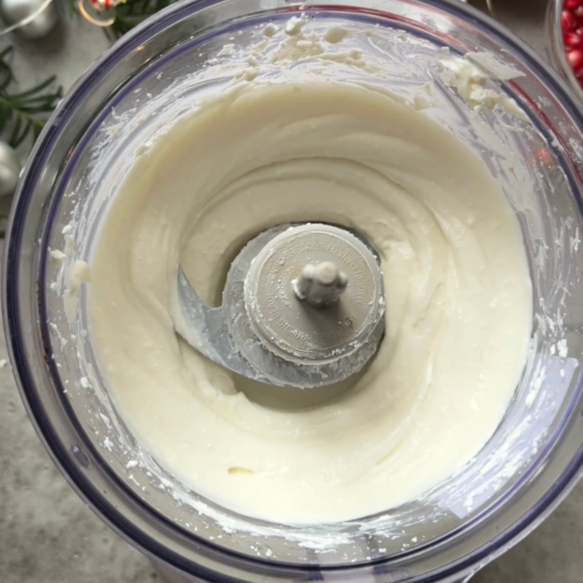 A food processor bowl containing creamy white mixture with a metal blade in the center, viewed from above.
