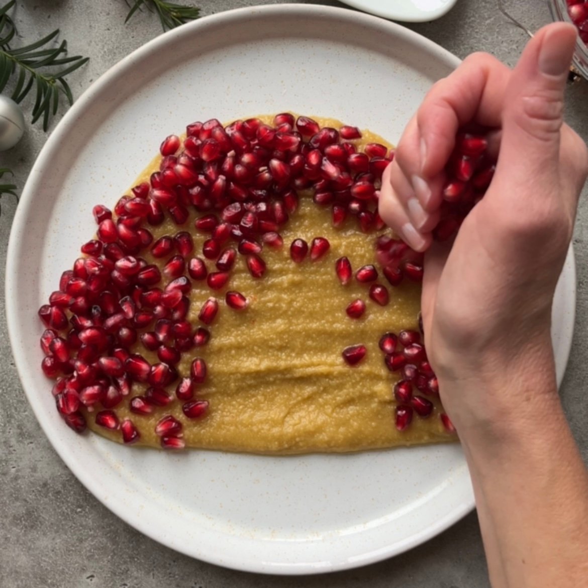 A hand sprinkles pomegranate seeds onto a smooth yellow spread on a round white plate.