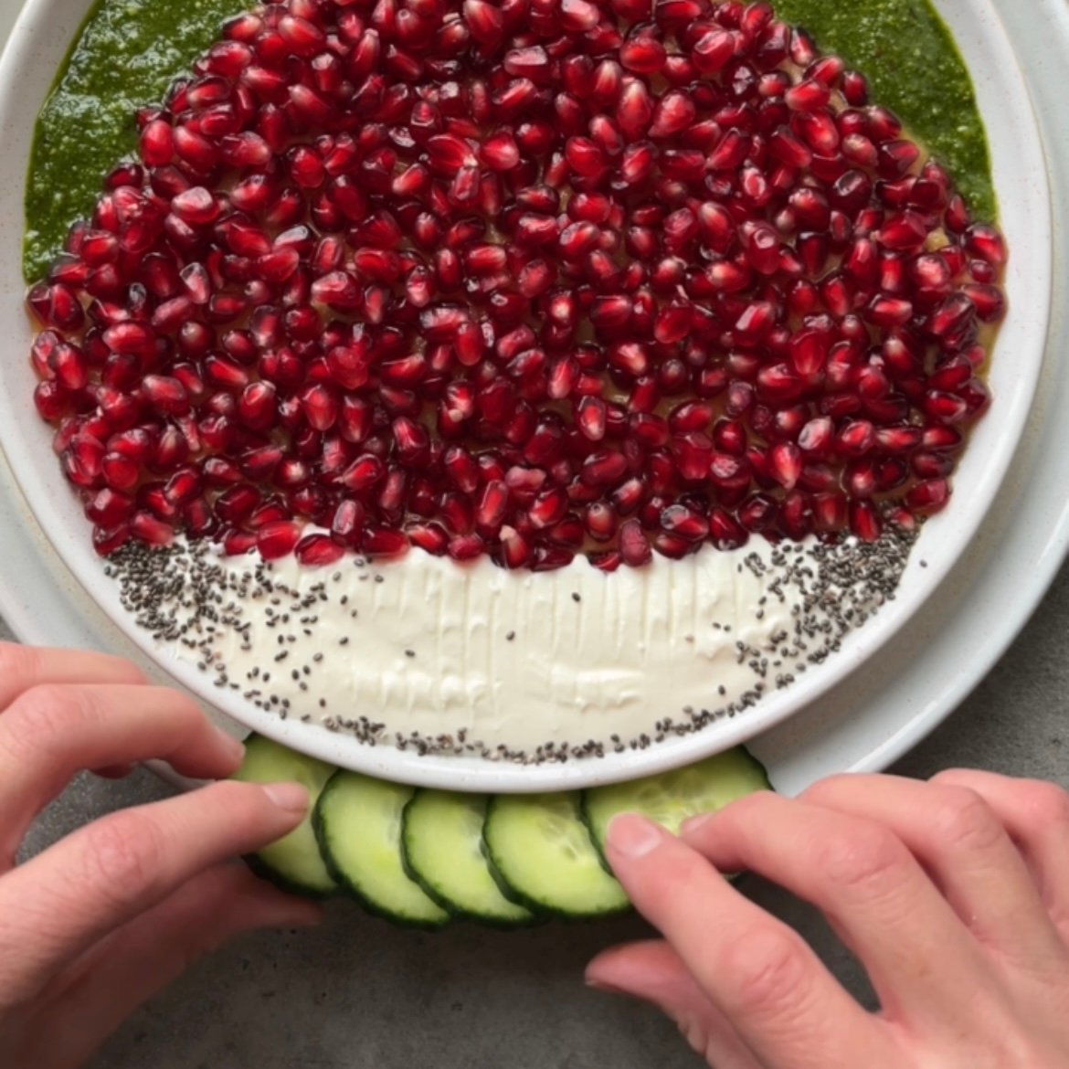 Two hands place cucumber slices around the trim of a plate, which features whipped feta and hummus topped with pomegranate seeds.