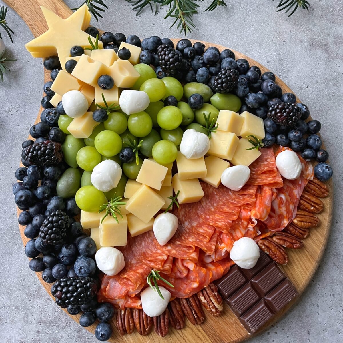 A charcuterie board arranged in the shape of a Christmas tree with cheese, grapes, berries, salami, pecans, chocolate, and mozzarella balls on a round wooden board.