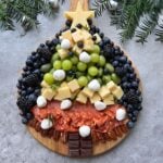 A Christmas tree-shaped charcuterie board with cheese, grapes, berries, mozzarella balls, salami, pecans, chocolate, and a star-shaped cheese topper on a wooden board.