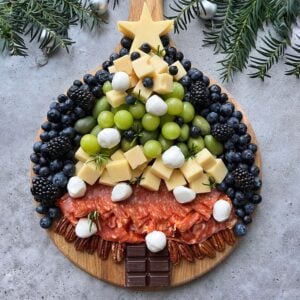A Christmas tree-shaped charcuterie board with cheese, grapes, berries, mozzarella balls, salami, pecans, chocolate, and a star-shaped cheese topper on a wooden board.