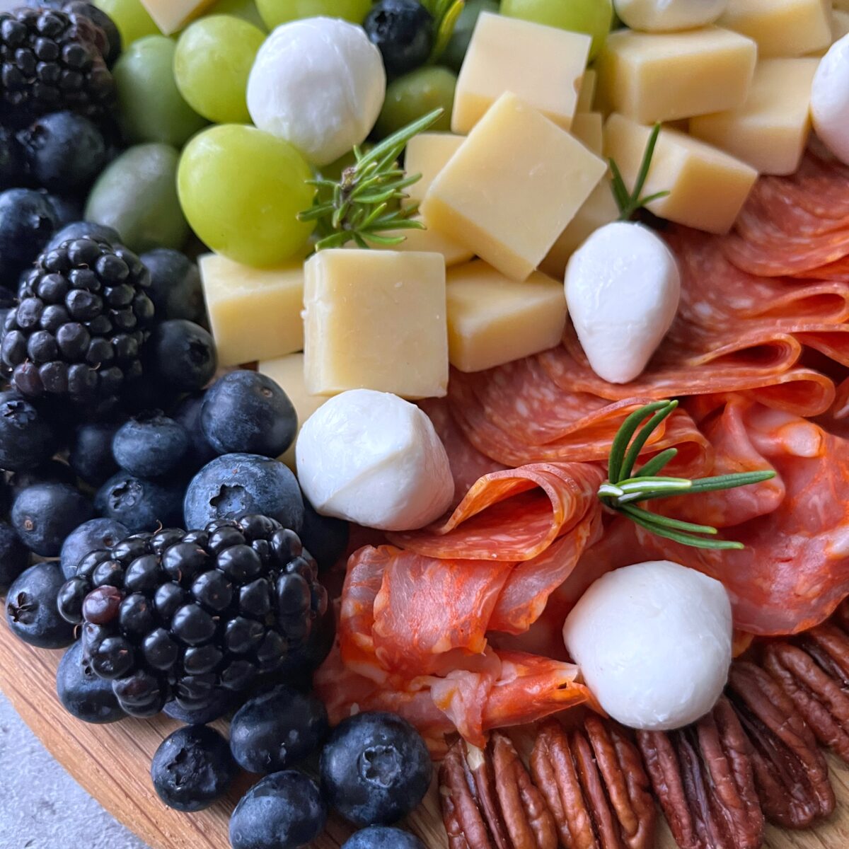 A charcuterie board with blackberries, blueberries, green grapes, cheese cubes, mozzarella balls, salami slices, and pecans, garnished with rosemary.