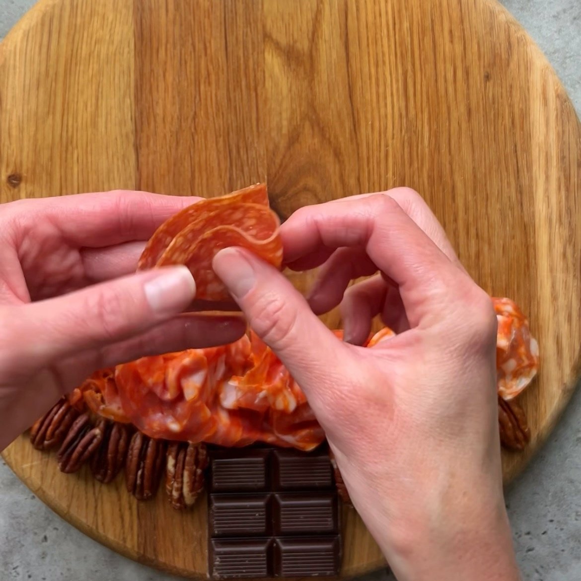 Hands folding a slice of cured meat over a wooden board arranged with slices of meat, pecans, and pieces of chocolate.