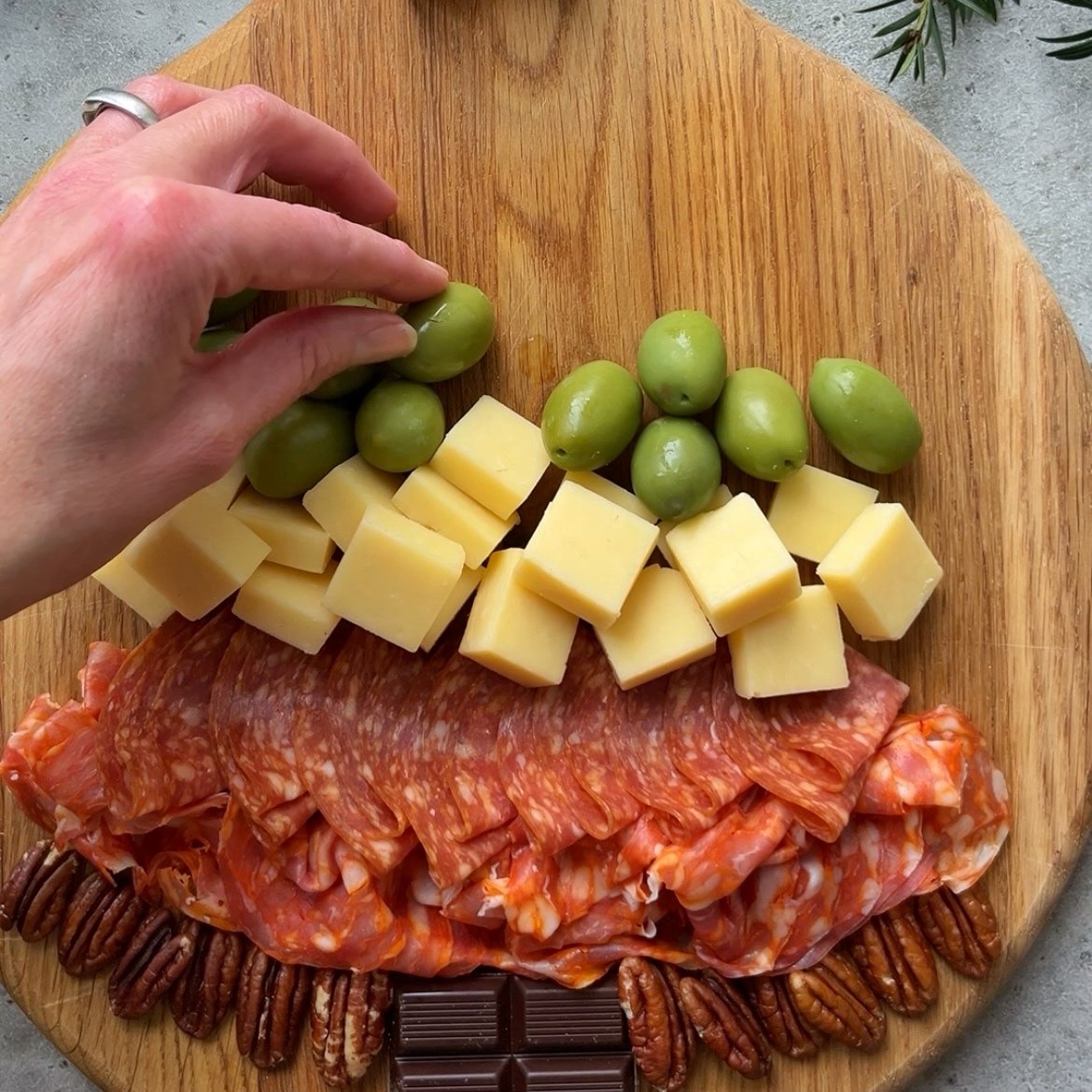 A hand arranging green olives on a wooden board with cheese cubes, sliced cured meats, pecans, and pieces of dark chocolate.