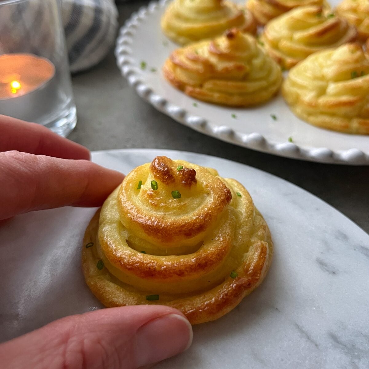 A hand picks up a piped, baked potato mound garnished with chopped herbs; more are arranged on a white plate in the background.