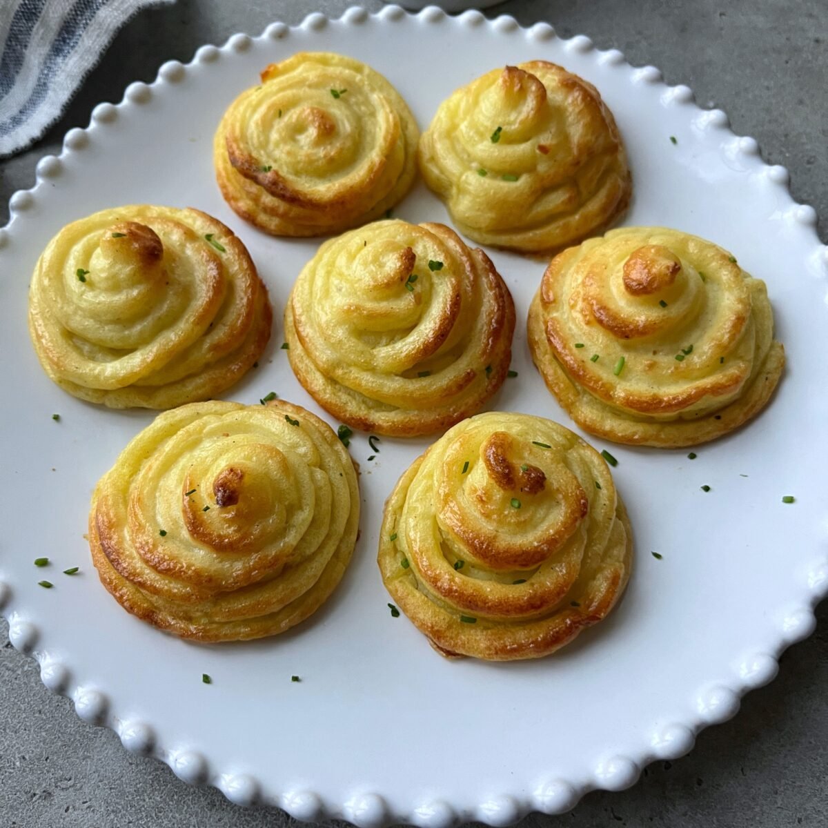 Seven piped and baked duchess potatoes are arranged on a white plate with a scalloped edge, garnished with finely chopped herbs.