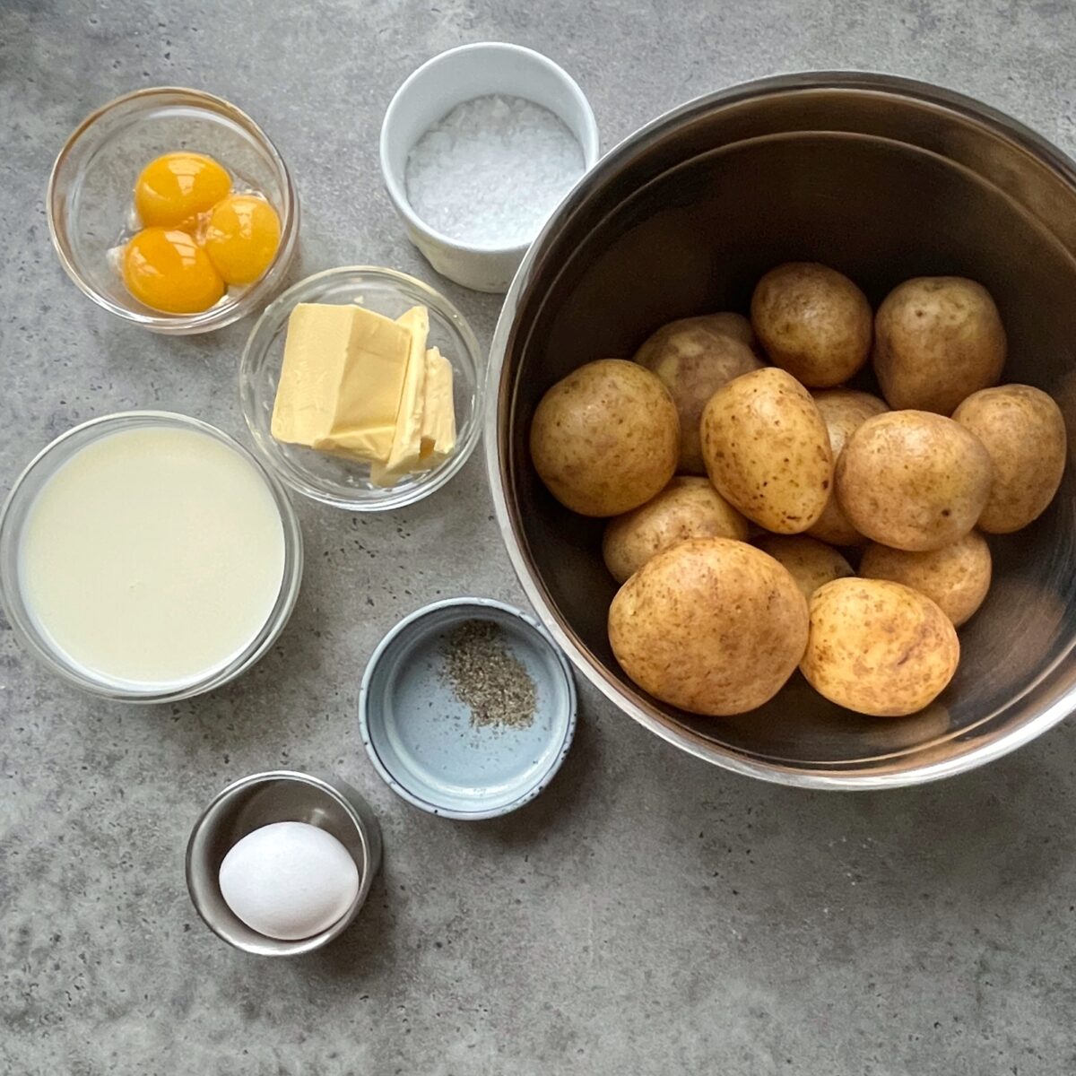 A metal bowl of whole potatoes beside small bowls containing egg yolks, butter, milk, salt, pepper, and a whole egg on a gray countertop.