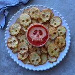 A plate of skull-shaped potato slices arranged around a bowl of dip with a skull face design made from sauce on top.