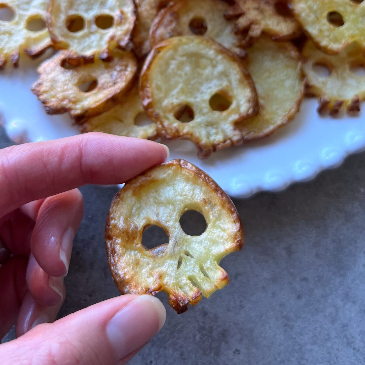 A hand holds a skull-shaped potato chip in front of a plate with more skull-shaped chips on it.