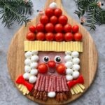 A festive food arrangement on a wooden board depicts Mrs Claus made from ham, cheese, cherry tomatoes, mozzarella, salami, pecans, and vegetables.