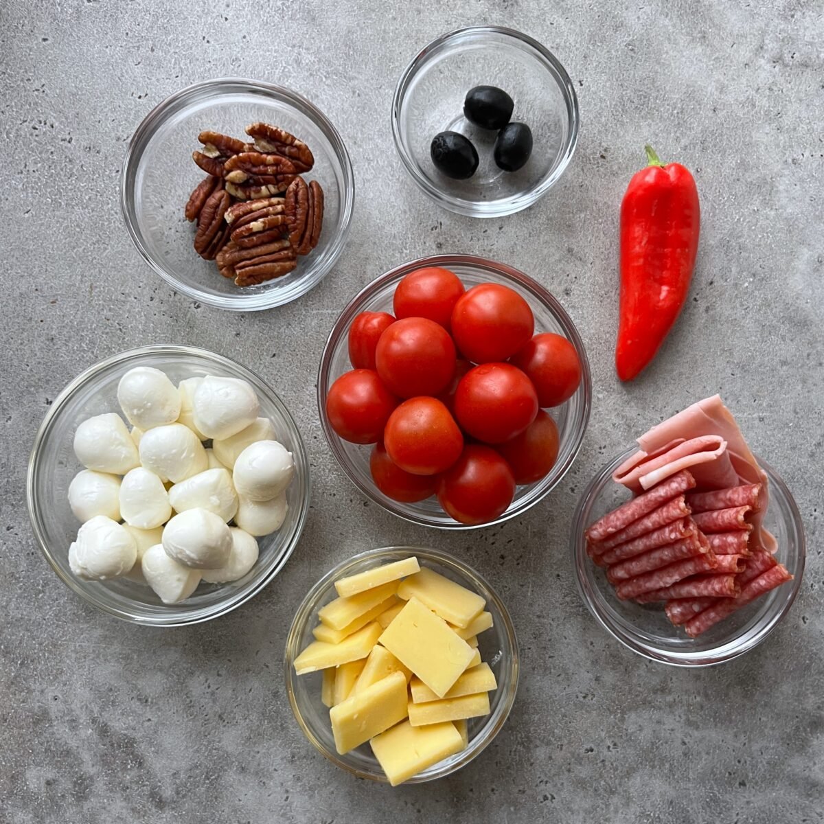 Seven small glass bowls containing pecans, black olives, cherry tomatoes, mozzarella balls, sliced cheddar, salami with ham, and a red bell pepper arranged on a gray surface.