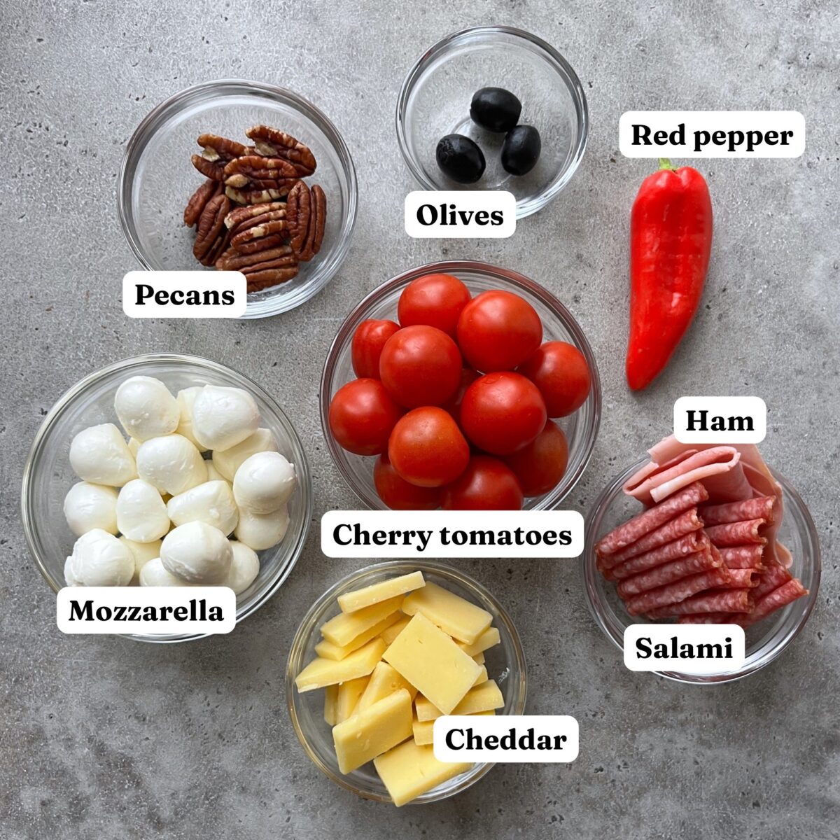 Six bowls and one red pepper on a grey surface contain pecans, black olives, cherry tomatoes, mozzarella balls, cheddar cheese, ham, and salami, each labeled accordingly.