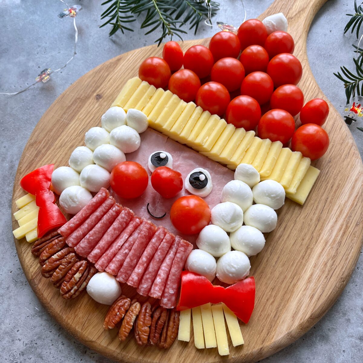 Side shot of charcuterie arranged as a Mrs. Santa face with cherry tomatoes, mozzarella, cheese, ham, salami, pecans, and olives on a wooden board with festive greenery nearby.