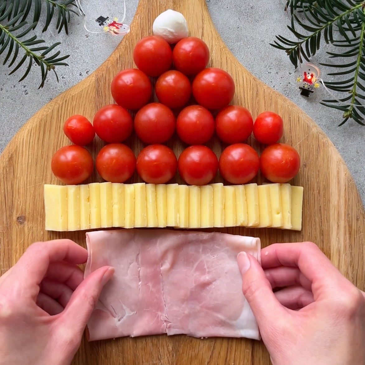 Two hands arranging a slice of ham on a chopping board at the base of a row of cheddar slices topped with a cherry tomato and mozzarella ball hat.