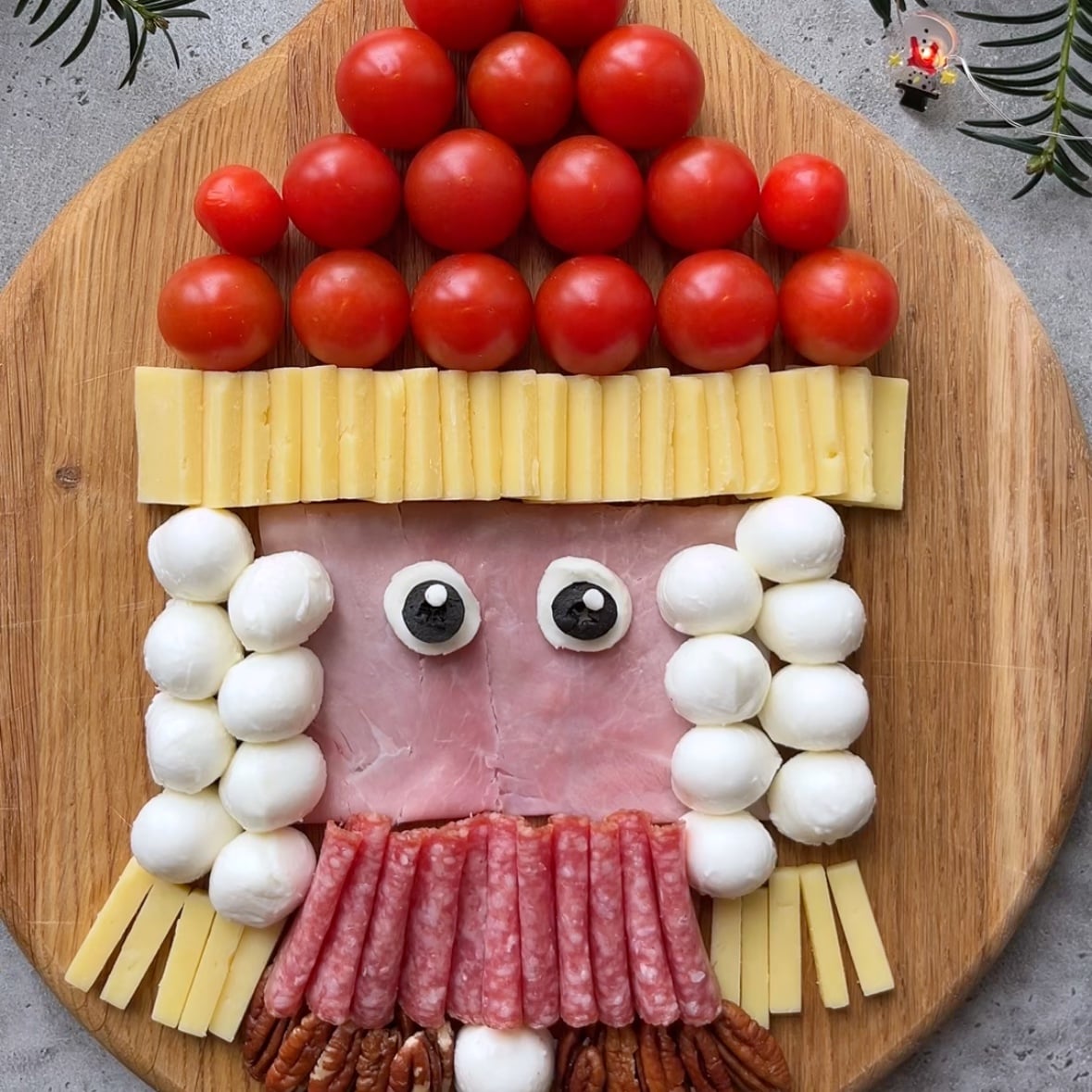 Holiday Charcuterie Board arranged as a face using cherry tomatoes, cheese, mozzarella balls, ham, olives, salami, and pecans on a wooden board, surrounded by festive greenery.
