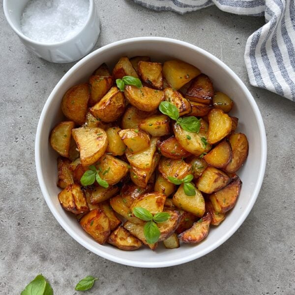 A white bowl filled with roasted potato chunks garnished with fresh basil leaves, placed on a gray surface near a small bowl of salt and a striped cloth.