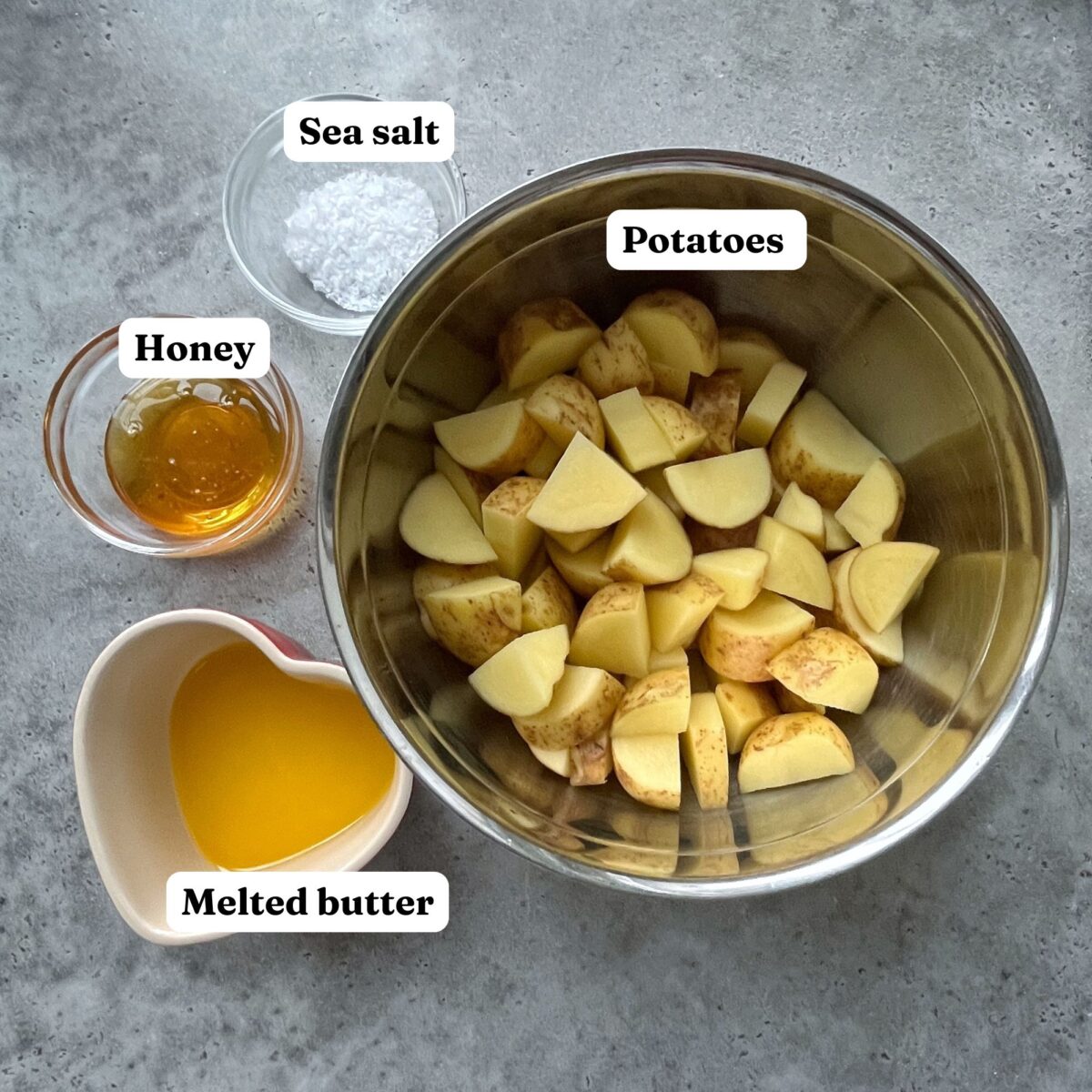 Chopped potatoes in a metal bowl next to small bowls of sea salt, honey, and melted butter on a gray surface.