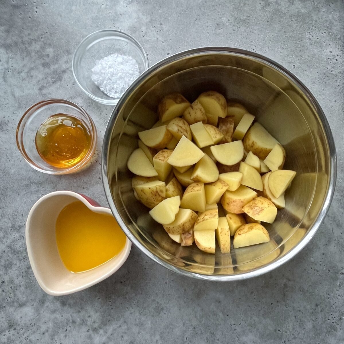A metal bowl of chopped potatoes, a small bowl of melted butter, a small bowl of honey, and a small bowl of salt on a gray surface.