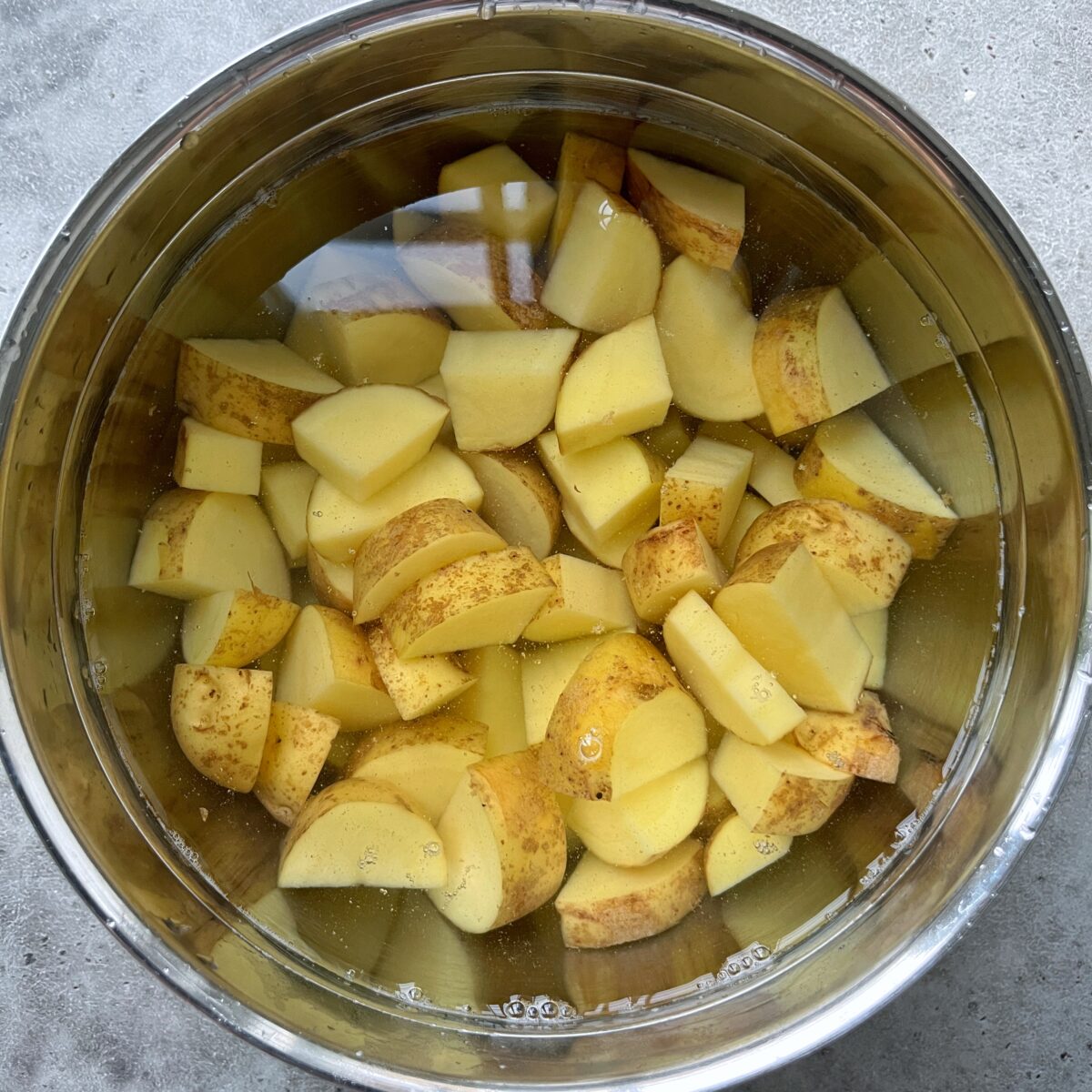 A metal bowl filled with water and chopped potatoes with skins on, soaking in preparation for delicious Honey Roasted Potatoes.