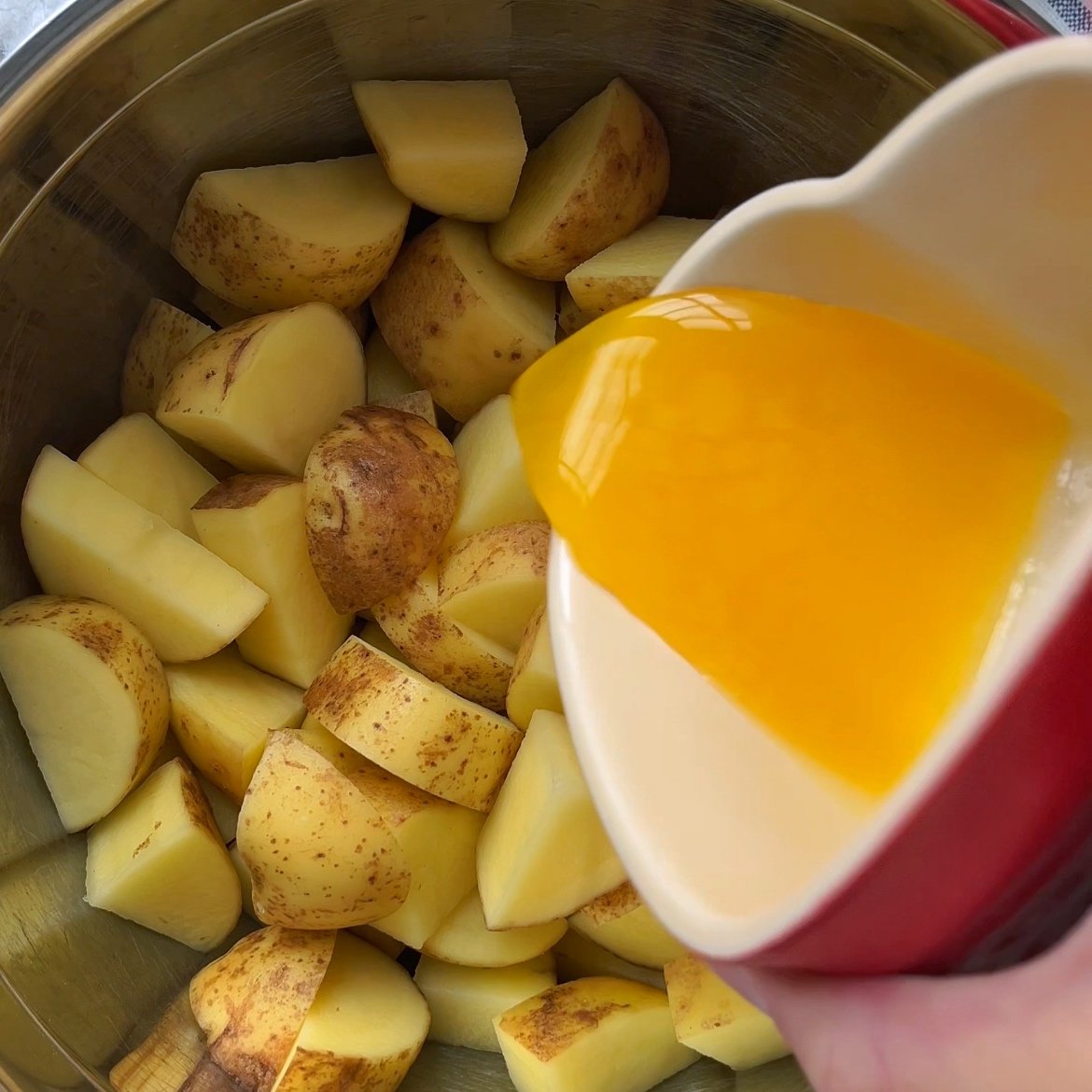 A hand pours melted butter from a red cup onto chopped, unpeeled potatoes in a metal pot, preparing them for delicious Honey Roasted Potatoes.