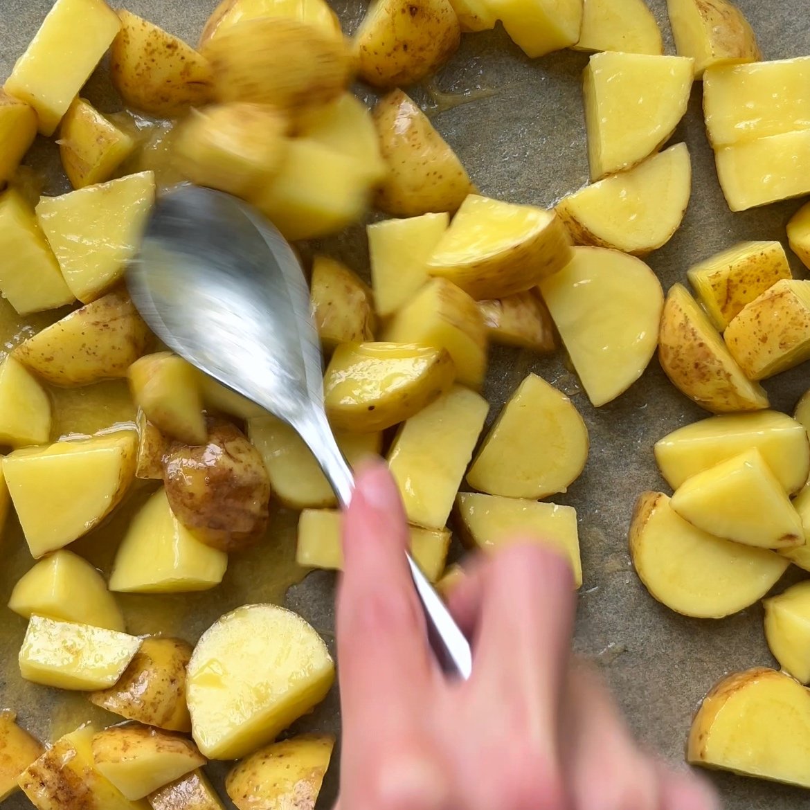 A hand uses a spoon to spread chopped, unpeeled potatoes coated in butter and honey onto a baking sheet, preparing them for honey roasted potatoes.