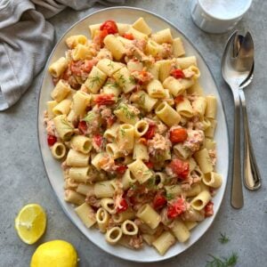 Oval platter of rigatoni pasta mixed with cherry tomatoes, salmon pieces, and fresh herbs, placed on a gray surface with a lemon, napkin, and utensils nearby.