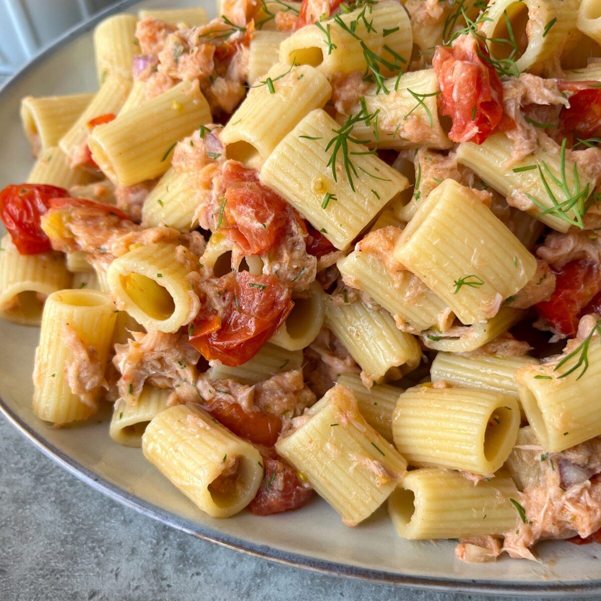 Close-up of rigatoni pasta mixed with cherry tomatoes, shredded salmon, and garnished with fresh dill on a light-colored plate.