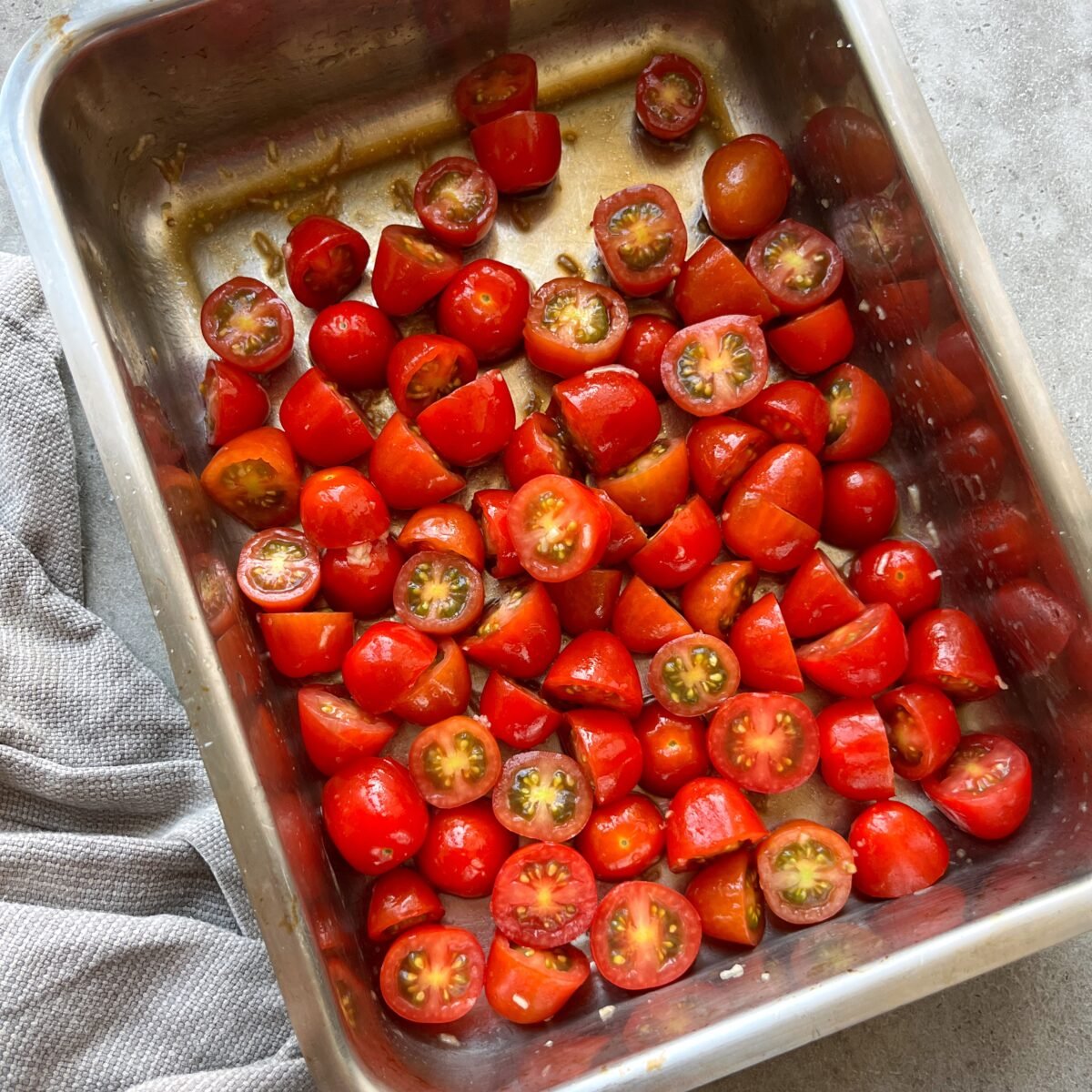 A metal tray filled with halved grape tomatoes, placed on a gray surface next to a textured cloth.