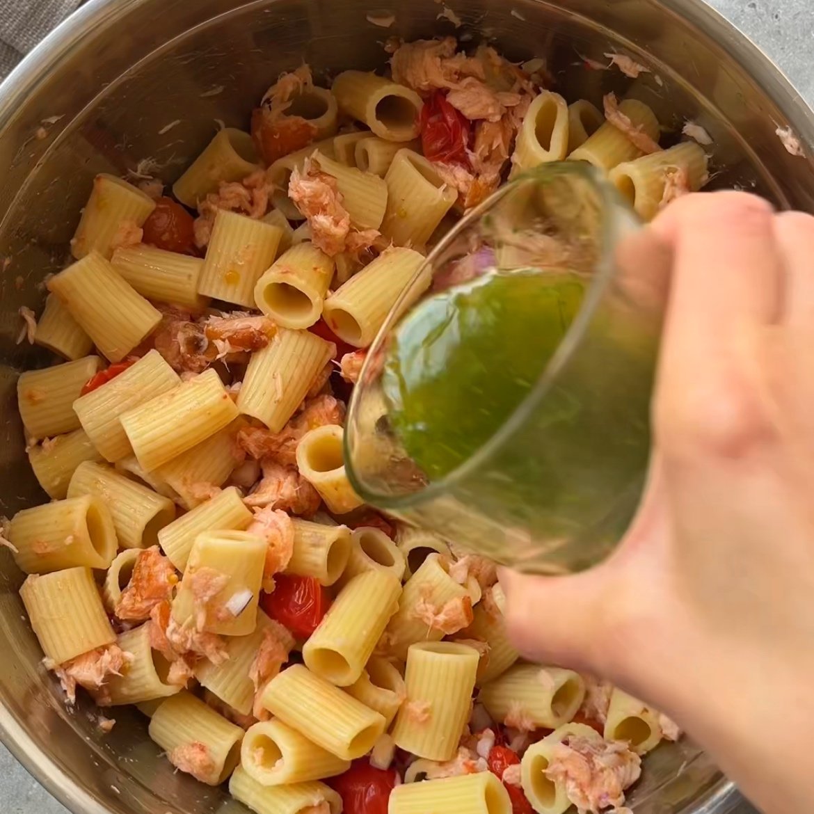 A hand pours green vinaigrette from a glass into a metal bowl containing pasta, baked grape tomatoes, red onion, and pieces of salmon.