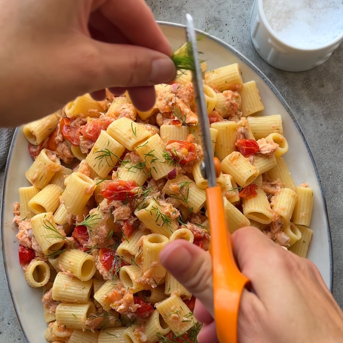 A person uses scissors to cut fresh dill over a plate of tubetti pasta with blistered grape tomatoes and hot smoked salmon. A small white bowl with sea salt sits nearby on a gray surface.