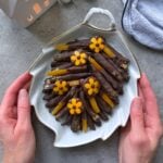 A person holds a leaf-shaped plate filled with chocolate-dipped candied orange peels, decorated with small yellow flower-shaped chocolates.